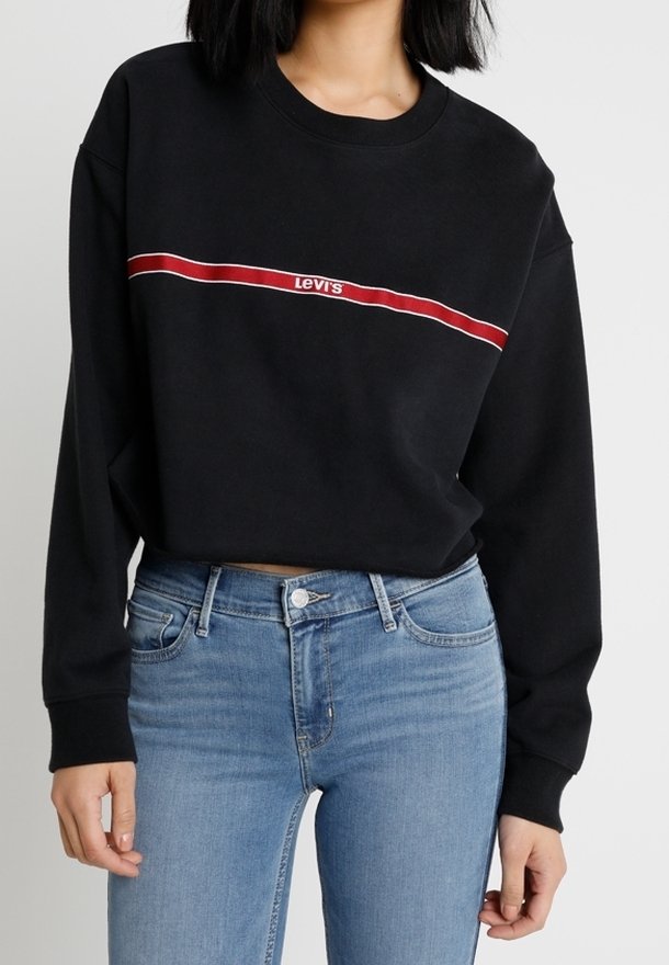 Femme portant un sweat-shirt court noir Levi's avec une bande rouge et blanche sur la poitrine et un jean taille haute bleu clair.
