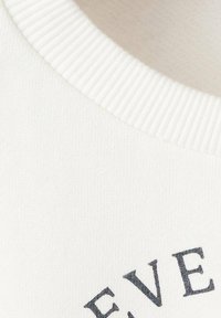 Tessuto di cotone bianco con scollatura a costine e testo grigio scuro in un carattere curvo, che mostra una morbida trama e una finitura liscia.