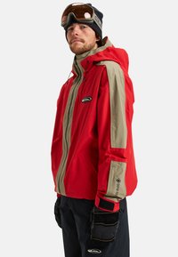 Man in een rode en beige Gore-Tex skijack, zwarte handschoenen, zwarte broek, een zwarte beanie en skibril op zijn voorhoofd.