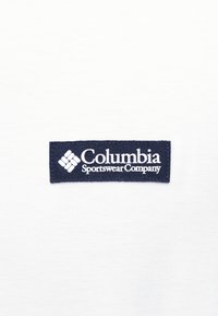 Ετικέτα με το λογότυπο της Columbia Sportswear Company με λευκό κείμενο και εικονίδιο σε σκούρο μπλε ύφασμα, τοποθετημένη σε λευκό ύφασμα.