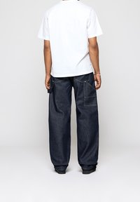 Valkoinen lyhythihainen t-paita yhdistettynä löysiin tummansinisiin farkkuihin, joissa on useita taskuja, näkyvät saumat ja rento siluetti.