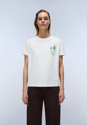 Donna che indossa una maglietta bianca con logo di una casa sull’albero verde e scritta sul petto, abbinata a pantaloni marrone scuro, in piedi davanti a uno sfondo semplice.