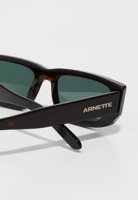 Hnedé slnečné okuliare s korytnačkovým vzorom a zelenými šošovkami, s obdĺžnikovým rámom a strieborným logom "ARNETTE" na nožičke.