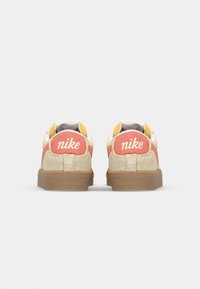 Nike tornacipő bézs velúrból készült felsőrésszel, rózsaszín és sárga díszítésekkel, fehér béléssel és barna gumitalppal. A márkázás a sarkokon látható.