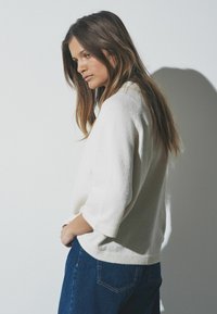 Bela, mehka pulover z dolgimi tremi četrtinami, v kombinaciji s temno modrimi kavbojkami. Obleka ima sproščen silhueto.