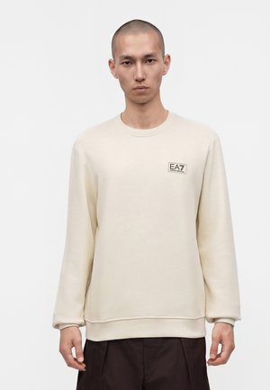 Mand med barberet hoved iført en creme sweatshirt med lille EA7 Emporio Armani-logo på brystet og mørke bukser, stående mod en hvid baggrund.