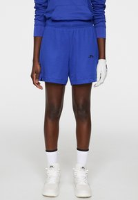 Siniset urheiluhousut pehmeästä materiaalista, joissa on joustava vyötärönauha ja pieni musta logo. Yhdistettynä valkoiseen käsineeseen ja lenkkareihin.