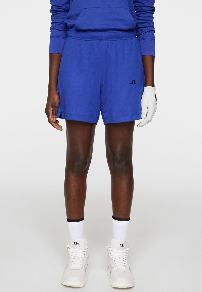 Siniset urheiluhousut pehmeästä materiaalista, joissa on joustava vyötärönauha ja pieni musta logo. Yhdistettynä valkoiseen käsineeseen ja lenkkareihin.