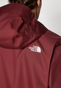 Chaqueta burdeos con capucha, hecha de una tela suave. Presenta un logo blanco en el hombro superior, que destaca un diseño distintivo.