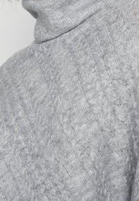Lysegrå strikket sweater med høj krave, der har et tekstureret kabelmønster og blødt, fluffy stof.