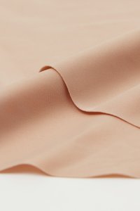 Glatter, hellbeiger Stoff mit feiner gewebter Struktur, sanft gefaltet, um weiche Kurven und leichte Schatten zu zeigen.