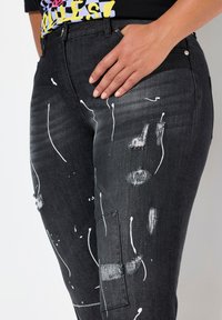 Jeans de mezclilla negros con salpicaduras de pintura blanca, parches desgastados, corte recto y detalle de bolsillo lateral, complementados con herrajes plateados.