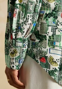 Cămașă cu mânecă lungă, având un imprimeu botanic colorat în nuanțe de verde, roșu și albastru pe un fundal alb, cu detalii vizibile ale nasturilor pe manșetă.