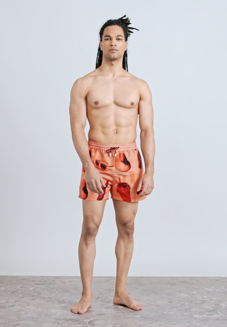 Homme musclé aux longues dreadlocks, debout pieds nus, portant un short de bain orange avec un motif de cocktail rouge, sur un fond gris uni.