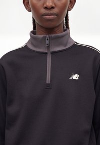 Άτομο με πλεξούδες που φοράει μαύρο φούτερ New Balance με φερμουάρ, με γκρι γιακά και λευκό λογότυπο στο στήθος.