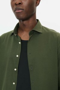 Close-up van een persoon met een open, olijfgroen, gestructureerd overhemd met knopen over een zwart shirt met ronde hals.