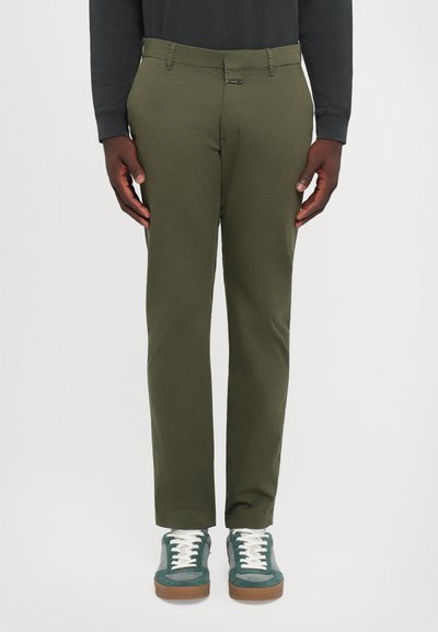 Homme portant un pantalon vert olive, des baskets teal et gris avec des lacets blancs, se tenant devant un fond clair uni.