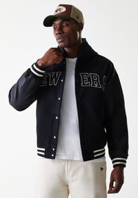 Navy varsity jacket with gray lettering, black leather sleeves, and striped cuffs. Worn over a white t-shirt, paired with light pants.