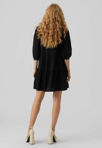 Robe noire à volants avec des manches bouffantes, fabriquée en tissu texturé. Le mannequin porte des sandales à talons blocs crème et a les cheveux lâches et ondulés.