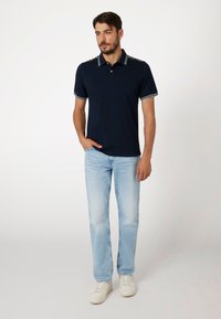 Marineblauwe poloshirt met contrasterende witte kraag en mouwtrim, gecombineerd met lichtblauwe jeans en witte sneakers. Eenvoudige, casual outfit.