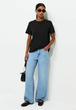 Kobieta ubrana w czarny t-shirt, jasnoniebieskie szerokie jeansy, czarne buty, kolczyki koła i czarne okulary przeciwsłoneczne, trzymająca małą czarną torebkę.