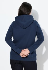Sudadera de color azul marino con capucha ajustable, puños y dobladillo acanalados. El material parece suave y texturizado. Se usa al revés con las manos entrelazadas por detrás.