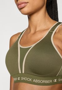 Αθλητικό σουτιέν σε ελαιί πράσινο με υφασμάτινη διακοσμητική υφή πλέγματος και μπεζ λεπτομέρειες. Διαθέτει φαρδιές τιράντες και ελαστική ταινία με λογότυπο για υποστήριξη.