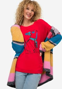 Rotes T-Shirt mit einem gedruckten blauen Grafikdesign, kombiniert mit einer mehrfarbigen, grob gestrickten Strickjacke mit gelben, pinken und blauen Streifen.