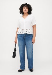 White short-sleeve blouse with lace trim, button front, and scalloped bottom; paired with blue straight-leg jeans and black handbag.