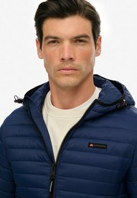 Młody mężczyzna z ciemnymi włosami w granatowej pikowanej kurtce z kapturem, noszący białą bluzę z okrągłym dekoltem, zwrócony twarzą do przodu z neutralnym wyrazem twarzy.