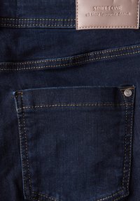 Tmavě modrá denimová tkanina s zadní kapsou, kontrastním prošíváním, kovovým nýtkem a béžovou nášivkou s nápisem "STREET ONE all time favourite denim."