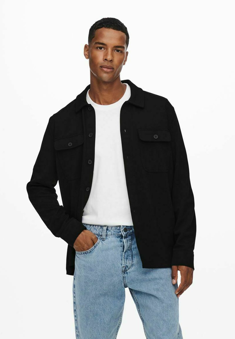 Camisa negra de botones con dos bolsillos en el pecho, llevada sobre una camiseta blanca, combinada con unos jeans de mezclilla azul claro. Tejido suave, corte relajado.