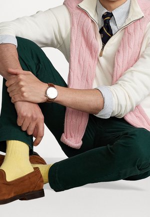 Mężczyzna siedzący po turecku, ubrany w zielone spodnie, żółte skarpetki, brązowe skórzane buty, biały sweter, różową kamizelkę z wzorem w warkocze, pasiastą koszulę i granatowy krawat.