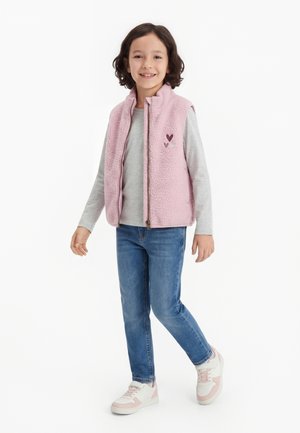 Barn med kort brunt hår har på seg en rosa fleecevest over en lys grå langermet skjorte, blå jeans og hvit-rosa joggesko, smilende mens han går.