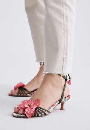 Pies con pantalones cortos color beige claro y sandalias slingback florales con decoraciones de flores rosas en los dedos.