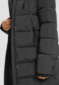 Roka pielāgo zīmi uz garā melnā, izšūta pūkainā jakā ar redzamām spiedpogu pogām un elastīgu piedurkni.