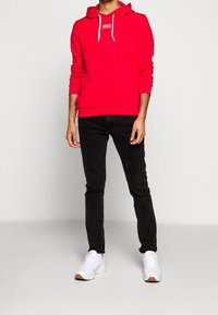 Sudadera roja con cordones blancos y logo, combinada con jeans ajustados negros y zapatillas blancas con suela de goma. Diseño simple y color vibrante.
