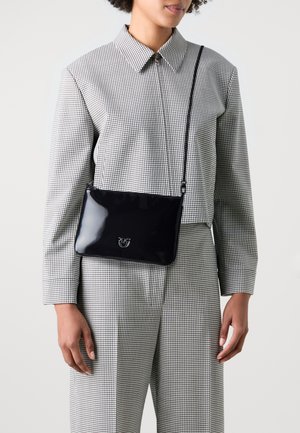 Borsa a tracolla in pelle verniciata nera con una texture liscia, caratterizzata da un sottile accento del logo. Il modello indossa una camicia a quadri e pantaloni.