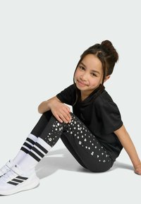 Black T-shirt, patterned black leggings with silver dots, and white sneakers with black stripes. The child sits with legs bent, smiling.