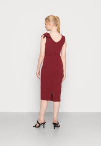 Vestido justo cor bordeaux com costas em V e laços nos ombros; com comprimento até ao joelho e uma fenda nas costas. Usado com sandálias pretas de salto alto sobre um pavimento claro.