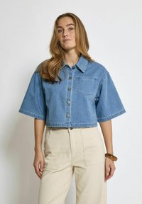 Himmelblaues, kurzes Denim-Hemd mit Knöpfen, kurzen Ärmeln und einer Brusttasche. Kombiniert mit hochgeschnittenen beigen Hosen.