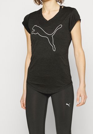 Femme portant un t-shirt noir Puma et des leggings avec des logos Puma blancs, debout devant un fond clair uni.