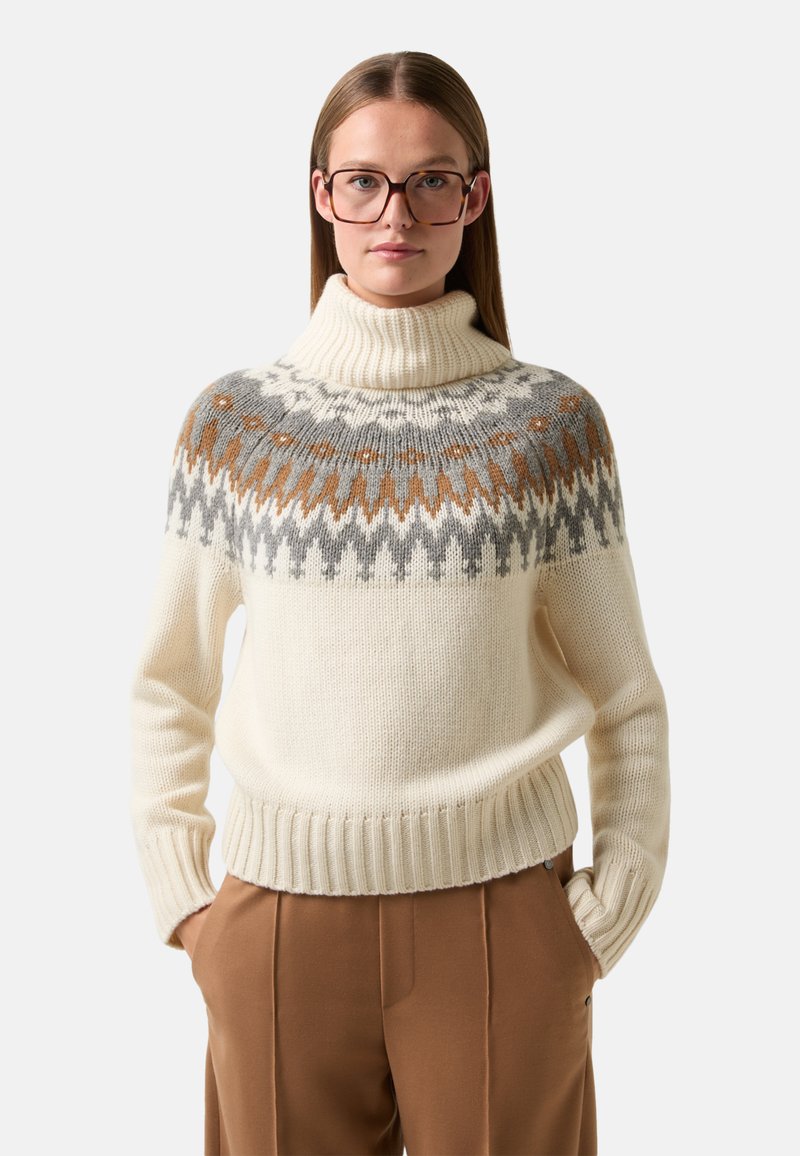 Ivoor gebreide trui met hoge kraag, voorzien van een patroontje in grijs en bruin op de schouders. Geribde manchetten en onderrand. Model draagt een bruine broek.