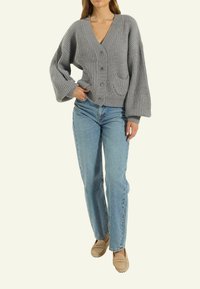 Grauer gerippten Cardigan mit V-Ausschnitt, Knopfverschluss und einer Tasche. Kombiniert mit hellblauen, geraden Jeans und beigen Slippern.