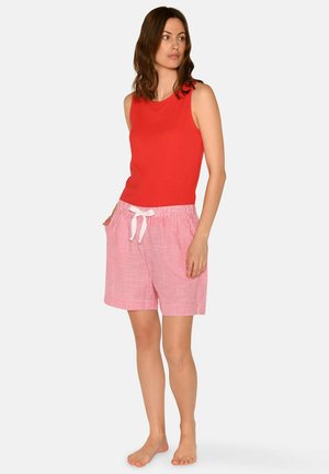 Kvinde står barfodet iført en ærmeløs rød top og lyserøde og hvide stribede shorts med snøre, hænderne i lommerne, kigger til siden.
