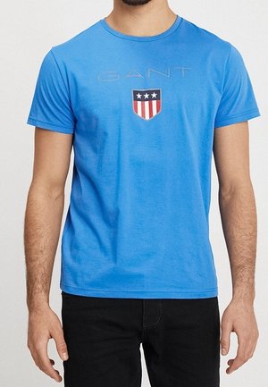 Blaues Baumwoll-T-Shirt mit rundem Ausschnitt, kurzen Ärmeln und einem mittigen grafischen Motiv mit einem Schilddesign in Rot, Weiß und Blau.