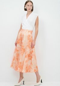 Weißes Wickeloberteil, kombiniert mit einem langen, fließenden Rock im orangefarbenen Blumenmuster, ergänzt durch cremefarbene Absatzschuhe mit einem schwarzen Akzent.
