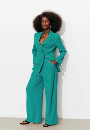 Femme aux cheveux afro naturels portant un blazer croisé vert sarcelle et un pantalon large, debout les mains dans les poches contre un mur blanc.