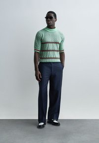 Mann mit Sonnenbrille, grünem kurzärmeligen Strickpullover mit Streifen, dunkelblauer Nadelstreifenhose und schwarz-weißen Mokassins, steht mit den Händen in den Taschen.