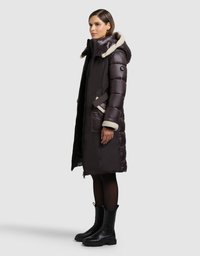 Długa, ciemnobrązowa puchowa kurtka z kapturem obszytym futrem, pionowym przeszyciami, bocznymi kieszeniami i zamkiem błyskawicznym, w zestawieniu z czarnymi botkami za kostkę.